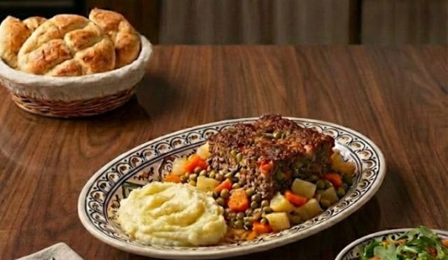 Fırın tepsisinde pişmiş, üzeri bezelye, havuç ve patates garnitürlü, nar gibi kızarmış sebzeli tepsi köfte sunumu.