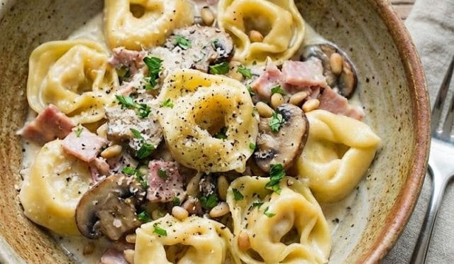 Beyaz bir tabakta sunulan üzeri taze baharatlar ve çam fıstığı ile süslenmiş yoğun krema soslu mantar ve salam dilimleriyle harmanlanmış nefis tortellini makarna.
