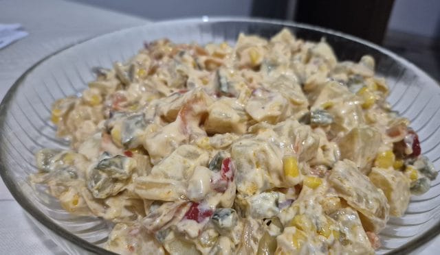 Karamel soğanlı, fırında pişmiş patatesli, turşu ve mısır içeren renkli bir patates salatası servis tabağında sunuluyor.