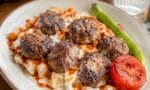 Balaban kebap yemeği, pide dilimleri üzerinde köfteler, yoğurt ve domates sosu ile servis ediliyor, yanında közlenmiş biber ve domates bulunuyor.