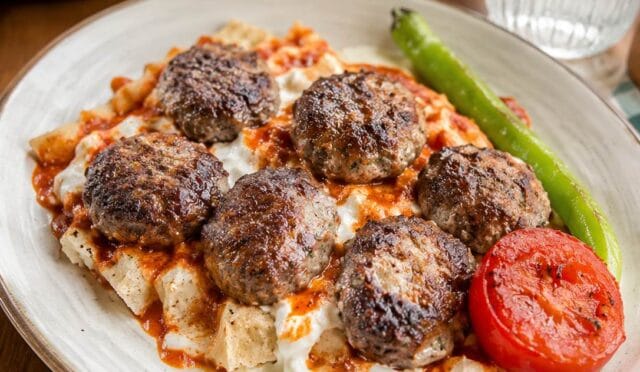 Balaban kebap yemeği, pide dilimleri üzerinde köfteler, yoğurt ve domates sosu ile servis ediliyor, yanında közlenmiş biber ve domates bulunuyor.