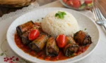 Domates sosu içinde pişmiş, üzerinde çeri domatesler ve ince kıyılmış maydanoz bulunan köfteli patlıcan sarmaları.